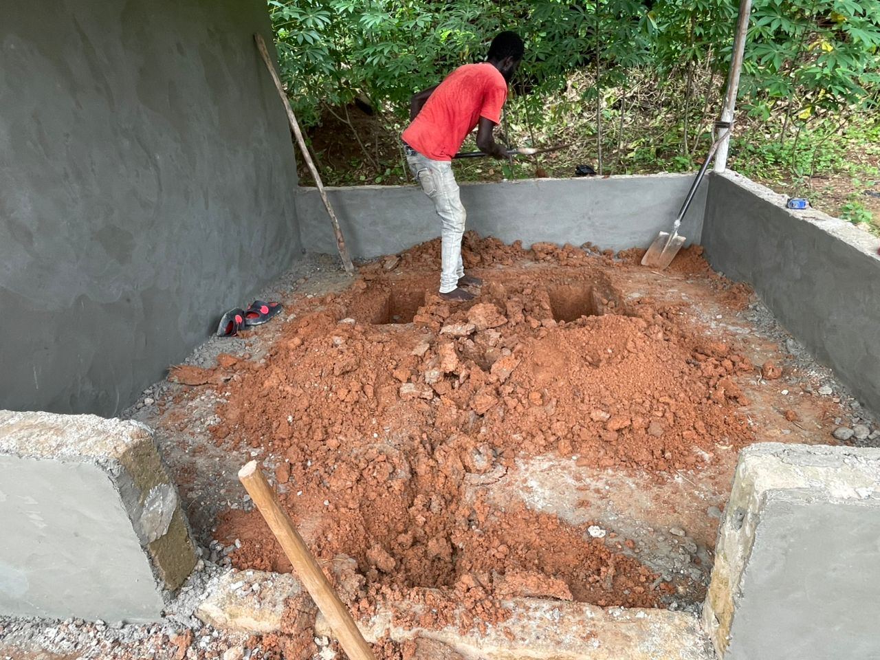 A worker digging soakaway pits inside a newly plastered block enclosure.